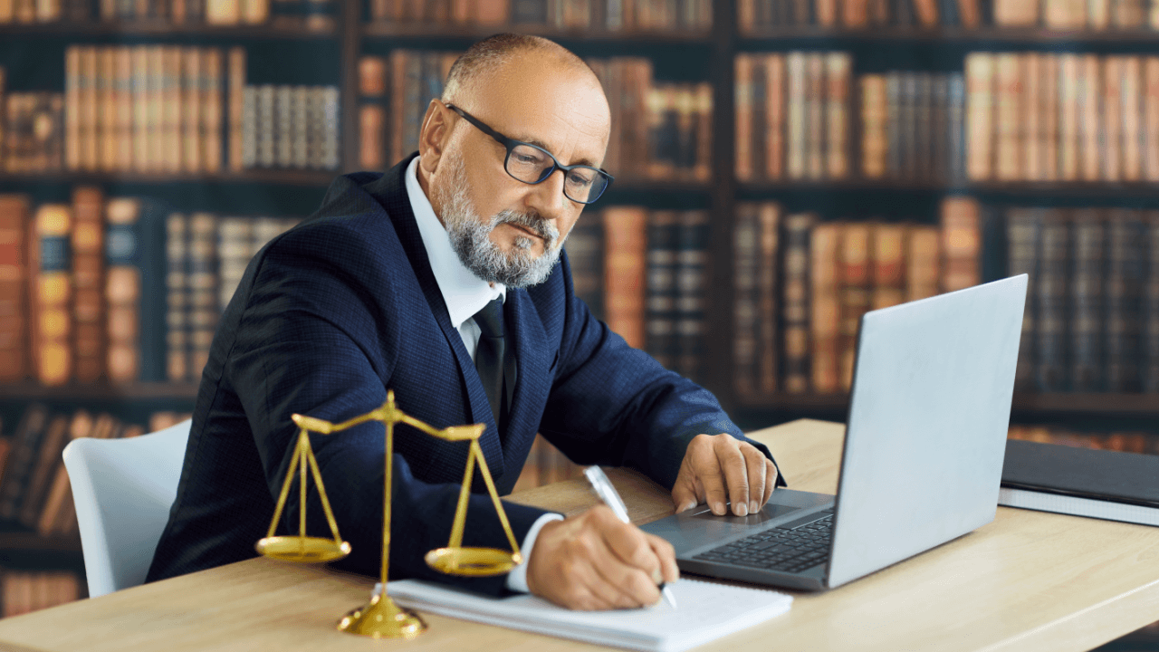 Employee rights lawyer writing case notes at desk with scales of justice and laptop in law library