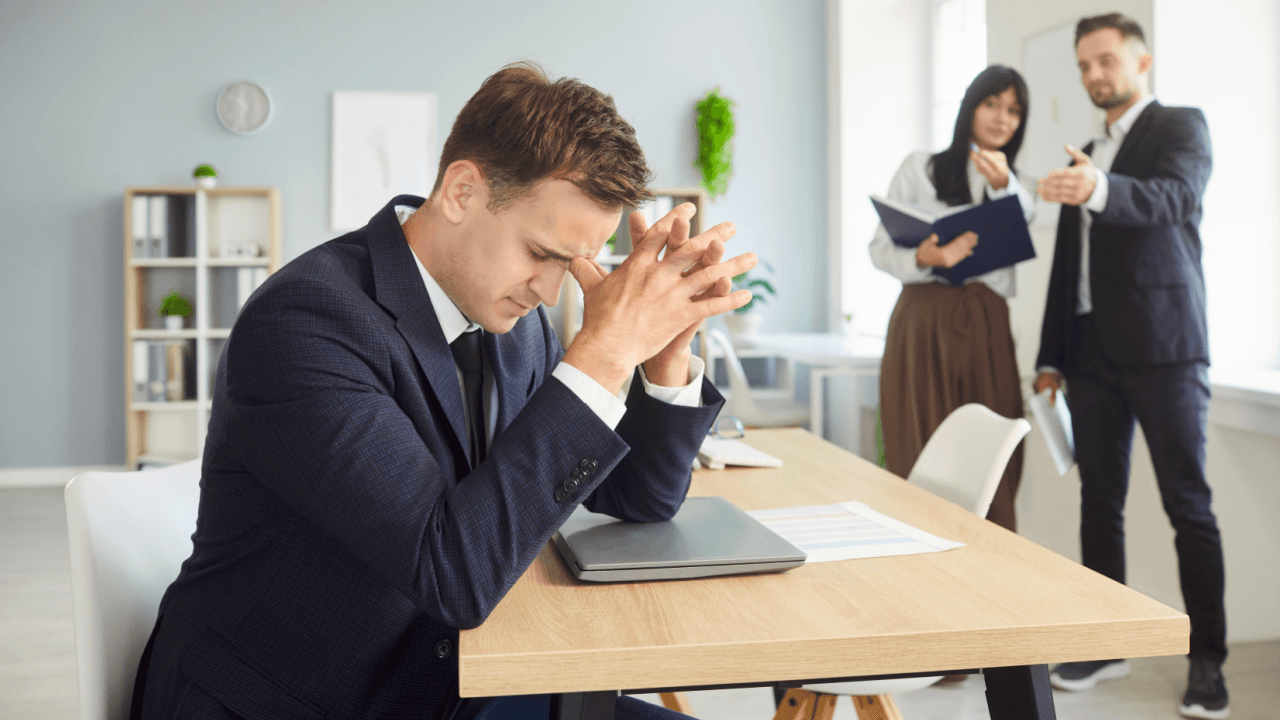 Stressed employee at desk understanding workplace bullying vs harassment from coworkers pointing behind him